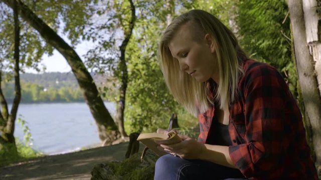Young Woman Sits In Peaceful Forest By Water, She Writes In Her Journal And Looks Around 
