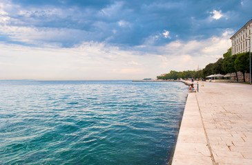Naklejka premium Stone tiles pavement near the sea with turquoise water against the background of small houses near the seaside in the distance and dramatic cloudy morning sky. Zadar, Croatia