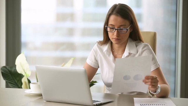 Annoyed busy businesswoman wearing glasses using laptop, doing paperwork, having problem with document, making error, miscalculating and retyping, finding discrepancies in graphs, missing deadline