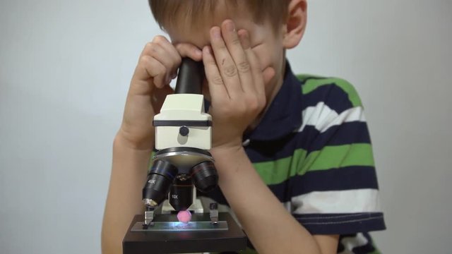 A boy in a striped T-shirt is studying a pink little ball through the eyepiece of a microscope against a white wall background, close-up shot