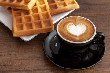 Coffee cup of latte art heart shape beside with some waffle on wooden background