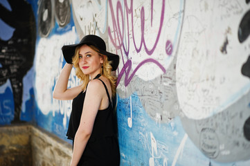 Naklejka premium Blonde woman on black dress, necklaces and hat against graffiti wall.