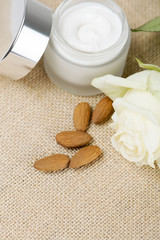 Close-up of a pot of skin cream next to almonds and a white rose on brown rustic fabric. Vertical shoot.