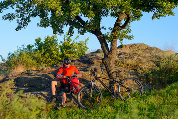 Cyclist in Red Jacket and Helmet Resting near Mountain Bike on Rocky Hill under Beautiful Green...