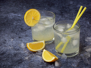 glasses of cold homemade lemonade with lemon slices, ice cubes and straws on dark background. Copy space.