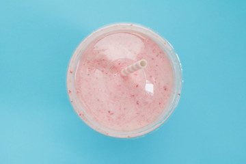 Top view of a strawberry smoothie in a plastic cup with a straw on a blue background. Pastel color. Summer & healthy lifestyle concept.