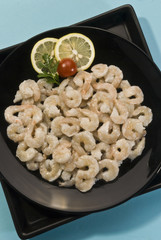 Raw baby shrimps with lemon and tomato on black plate on blue background