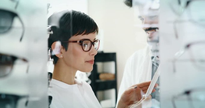 Beautiful woman with optician trying eyeglasses at optics store