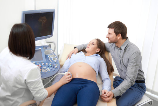 Brunette female doctor in white, looking at screen,diagnostic and screening stomach of  pregnant woman using tool and gel. Mom looking at picture of baby. Husband carrying about wife at ultrasound.