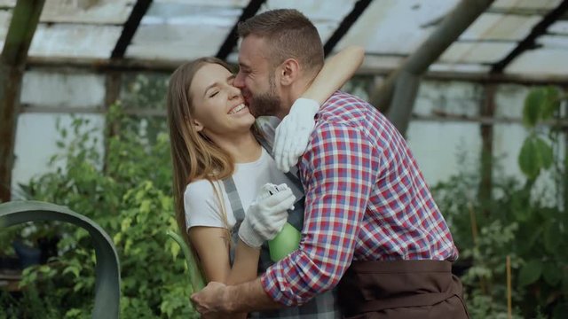 Happy young florist couple in apron have fun while working in greenhouse. Laughing woman spray water in husband face and embrace and kiss him