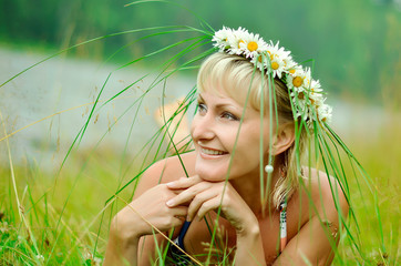 A young girl in a wreath of chamomiles lies on a green meadow. Wide, sincere smile