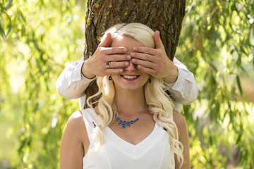 Couple in love. Man covered eyes of smiling blonde woman by his hands in park. Young people resting in nature, girl leaned against a ash tree,  natural environment background