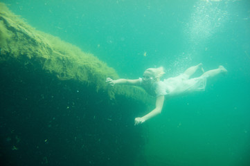 A floating woman. Underwater portrait. Girl in white dress swimming in the lake. Green marine plants and water