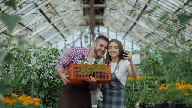 Cheerful loving couple gardeners taking selfie picture on smartphone camera and kissing while working in greenhouse