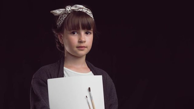 Portrait of young artist painter girl smiling to camera before black background, holding brushes and album