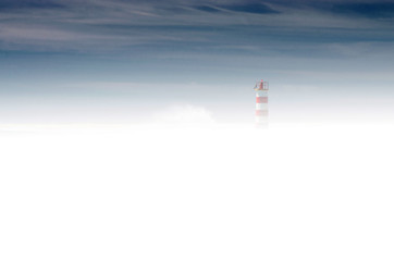 Sylwetka latarni morskiej wyłaniająca się spoza gęstej mgły. Granatowe niebo, gęsta mleczna mgła, drobna sylwetka latarni po prawej stronie.  - obrazy, fototapety, plakaty