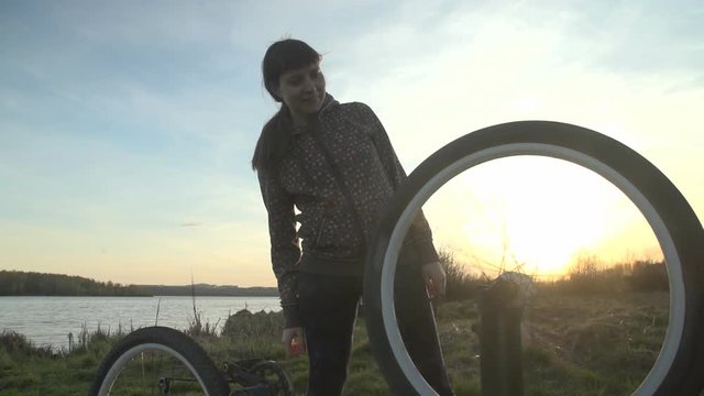 A young woman in a spotted sweatshirt checks her bicycle's wheel for damage on a summer evening near a pond in the rays of the setting sun.