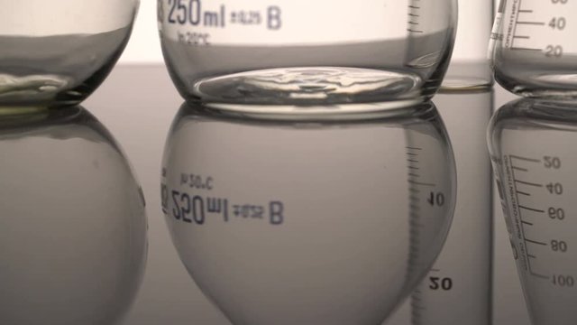 Tracking close-up shot of empty chemical glass flasks and test tube reflecting on the surface against white background.