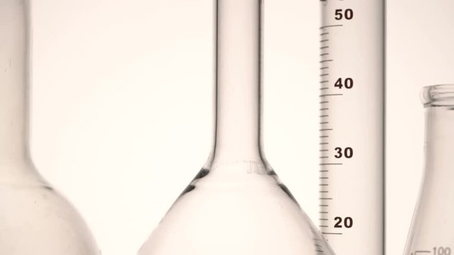 Tracking close-up shot of empty chemical glass flasks and test tube against white background.