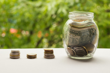Glass jar with money and stack of coins against background of greenery.