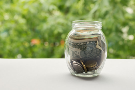 Glass jar with money and coins against background of greenery