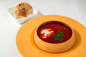 A delicious red soup with sour cream and herbs, in a dark yellow plate, and bread rolls, on a white tablecloth. Horizontal frame