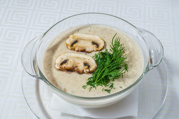 Delicious white soup with mushrooms and herbs, in a transparent plate, on a white tablecloth. Horizontal frame