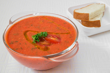 Delicious red soup with herbs in a transparent plate, and bread on a white tablecloth. Horizontal frame