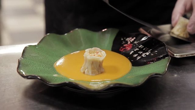 Chef Oriental restaurant puts on a plate dim sama with crab. He puts 4 Chinese dumplings on a green saucer with hieroglyphics, in which yellow bisque sauce is poured. The chef prepared a light meal