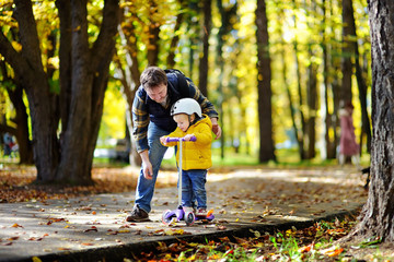 Middle age father showing his toddler son how to ride a scooter in a autumn park