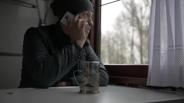 A young man goes on a train, answers a call, makes a call on a white smartphone, puts a smartphone on a table and takes a glass of tea, drinks