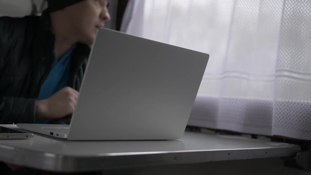 The young man goes by train in a gray cap and gray jacket, sits down at a table and talks on a video call of a laptop, laughs, gesticulates. Trees grow outside the window