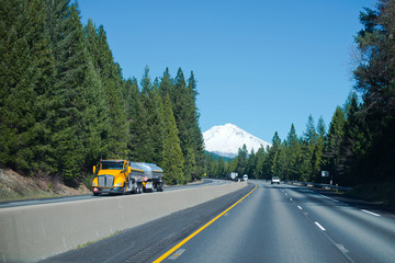 Chrome yellow semi truck with shiny stainless steel tanks at turn of scenic highway with firs and snowy mountain