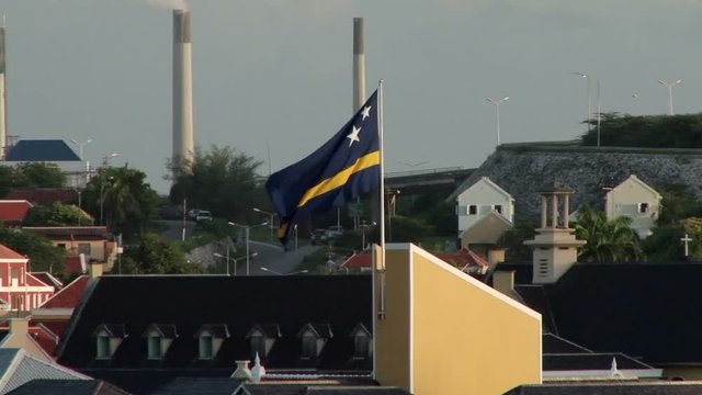 View from a ship as the camera tracks the flag of Curacao. Large industrial chimneys are in the background 