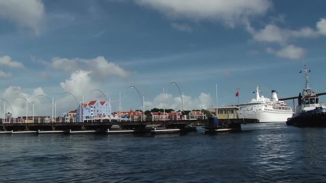 The Queen Emma Bridge begins to open across St Annes Bay in Willemstad, Curacao. An older cruise ship can be seen in the background and a tug boat is sailing past