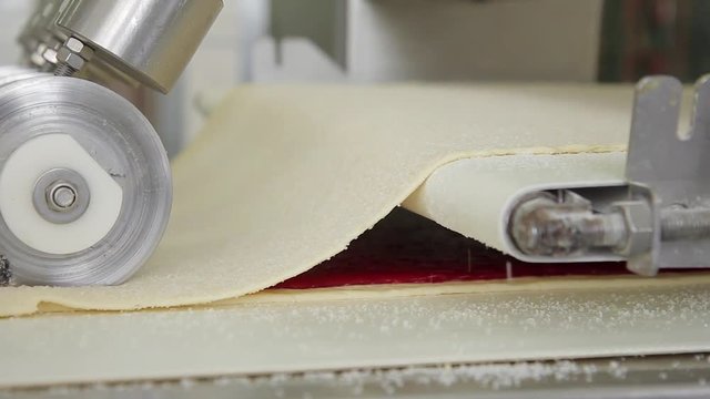 Part of process of cooking strudel, puff on conveyor in bakery factory. There are two levels of pastry moving on assembly line. Lower covered with thin smooth layer of dough with red jam on it