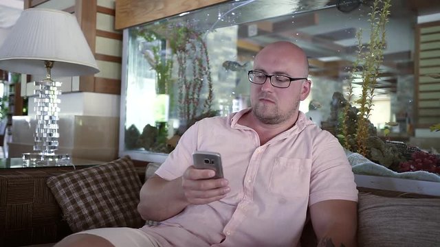 A bald man with glasses sits in a hotel on the sofa next to a large marine aquarium with fish and live algae. Holding a phone and taking pictures of themselves fooling around.