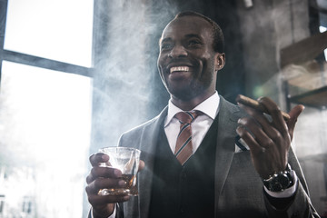 Smiling african american businessman holding glass with whiskey and smoking cigar