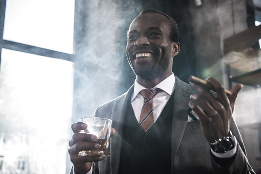 Smiling African American Businessman Holding Glass With Whiskey And Smoking Cigar