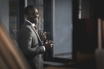 smiling african american businessman in suit with glass of whiskey smoking cigar
