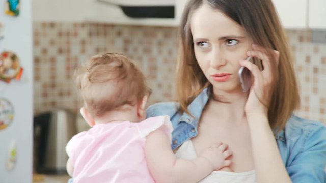 A beautiful and young mother is holding her little baby in her arms while talking on the phone. Modern mom holds a daughter in her arms and uses a mobile phone. Young woman with baby and phone.