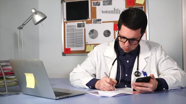 Medical assistant working at workplace with smartphone and medical forms and papers. He writes down the information.
