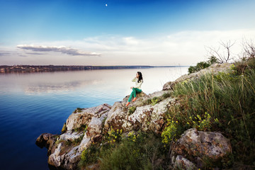 Fototapeta premium Young woman sits on rock and enjoys view of river at sunset