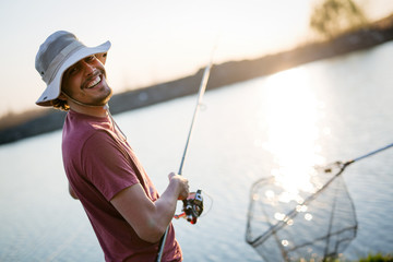 Young man fishing on a lake at sunset and enjoying hobby - obrazy, fototapety, plakaty