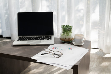 Obraz na płótnie Canvas office table with blank screen on laptop, coffee cup, garden plant pot and statistic report paper ob blurry drape at living room, concept of business lifestyle.