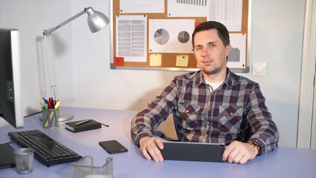 The young manager of the company sits at the desk where his devices are located, namely a tablet, a mobile phone, a notebook for recording important meetings and a keyboard from a computer