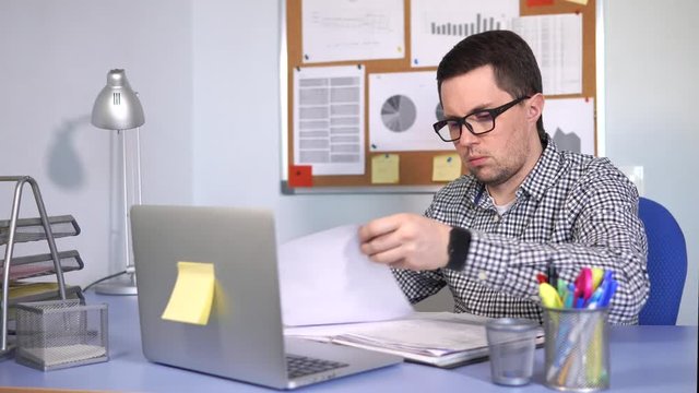 A busy man, similar to a manager, fills in the reporting documentation for his firm. A person with poor eyesight makes a marking pen on paper, on his desktop is a laptop