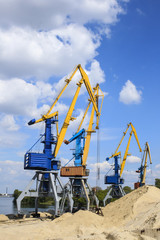 Fototapeta na wymiar Cranes in the river port on a blue sky background near the sand mound