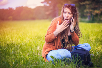 Shocked young woman receiving surprising news over her phone, sitting in the green grass in the nature on a bright sunny day