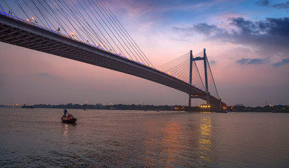 Fototapeta na wymiar Vidyasagar Seu (bridge) at twilight with a wooden boat on the river. The cable bridge connects Kolkata with Howrah district.